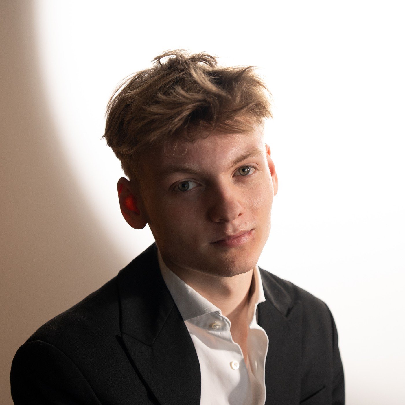 Simonas Vakaris Savickas in a dark blazer and white shirt, lit against a bright spotlight on a wall.