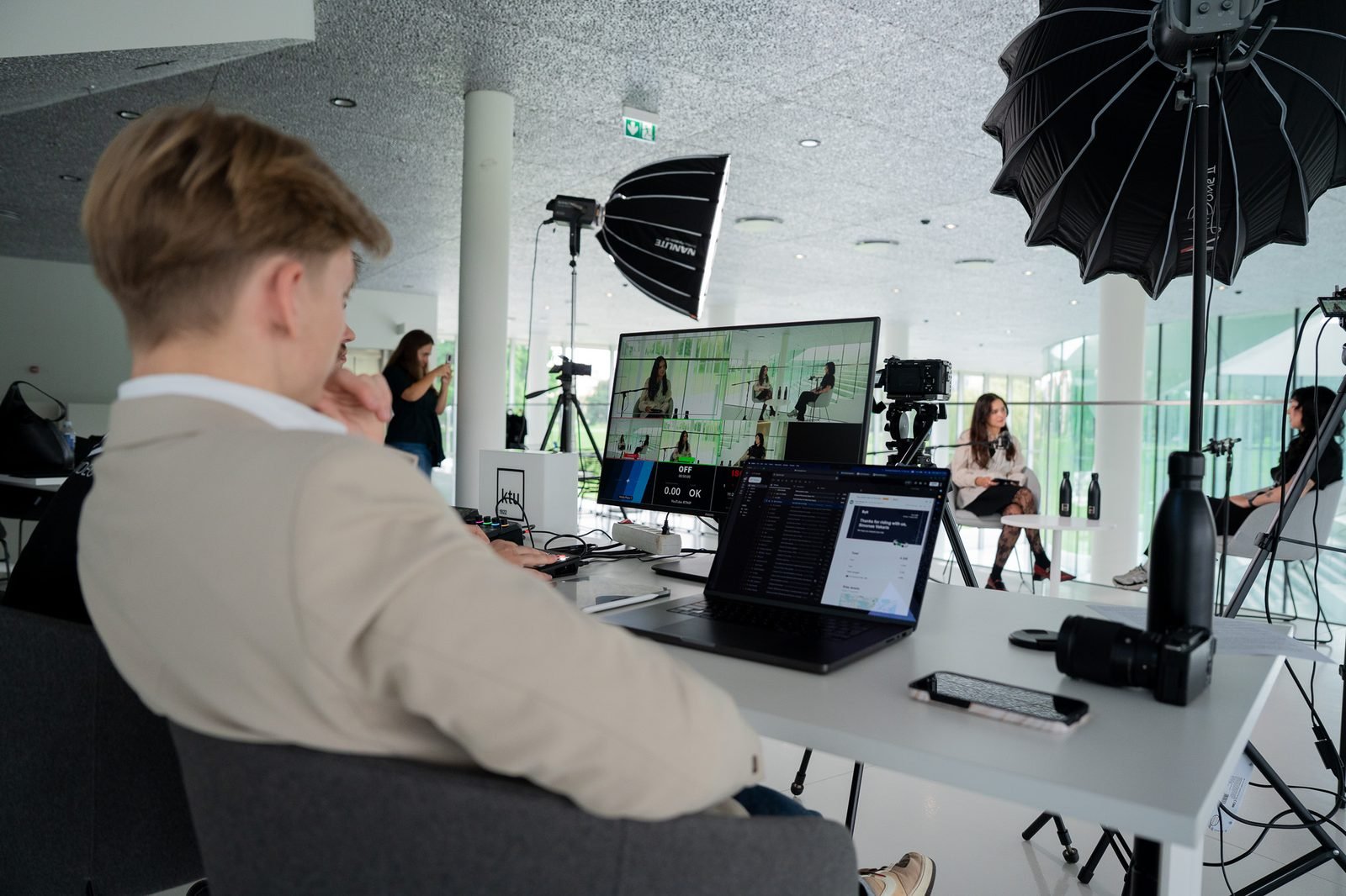 Simonas, in a beige blazer and seen from behind, watches the multicam monitor at the production desk of a live interview set. A crew member films in the background while two guests talk under softbox lights.