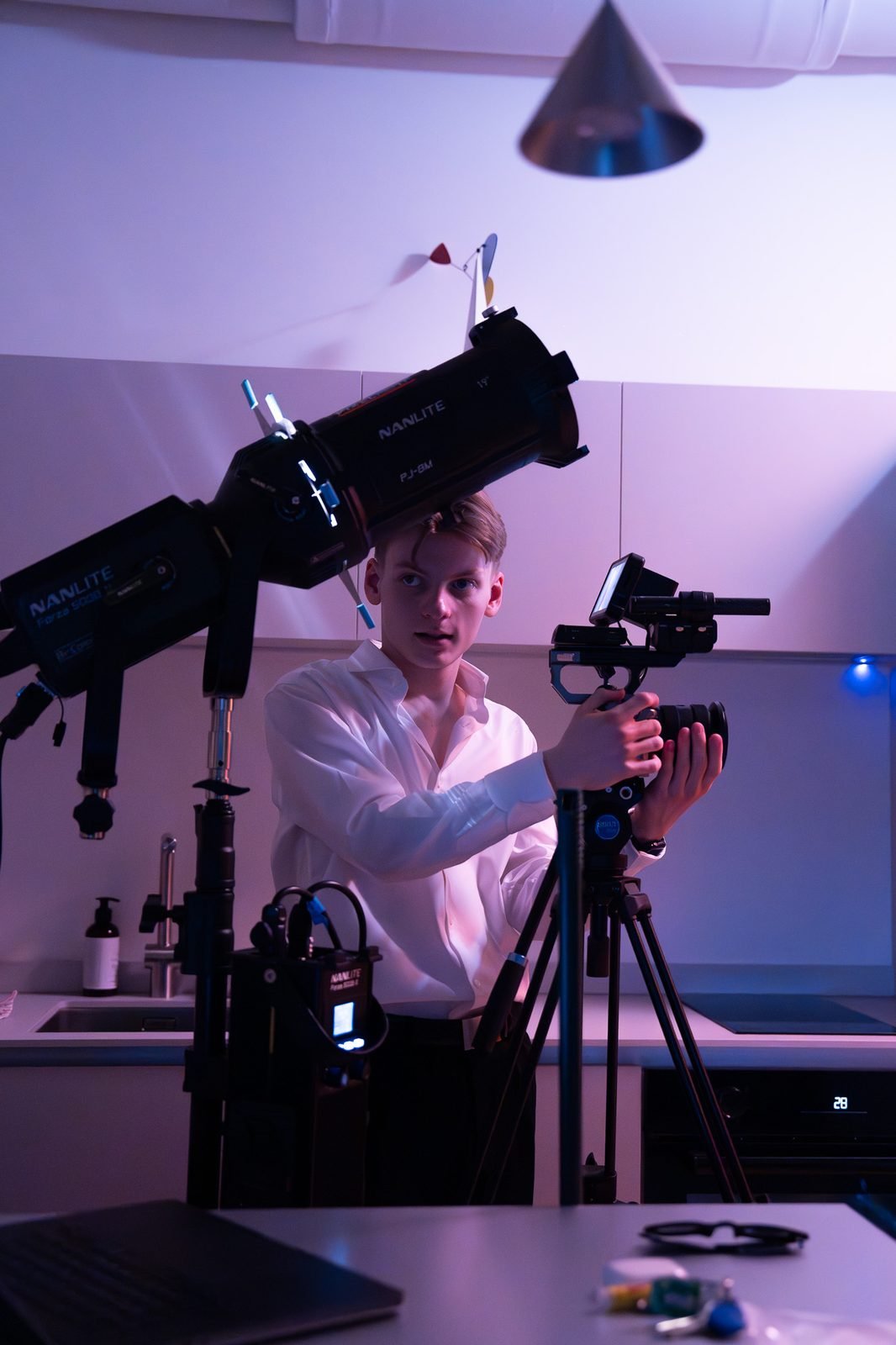Simonas alone on a small set, adjusting a camera on a tripod beside a spotlight, in purple light.