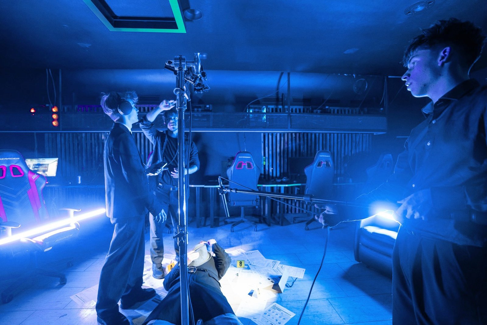 A crew in blue light sets up a light stand and boom around a scene, with Simonas among them on the left.
