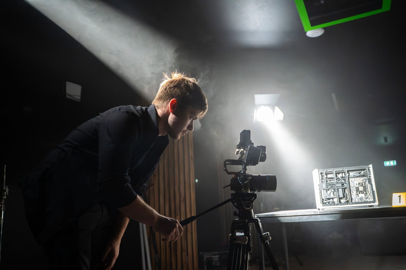 A camera operator leans in to adjust a camera on a tripod, beside a spotlight cutting through the haze.