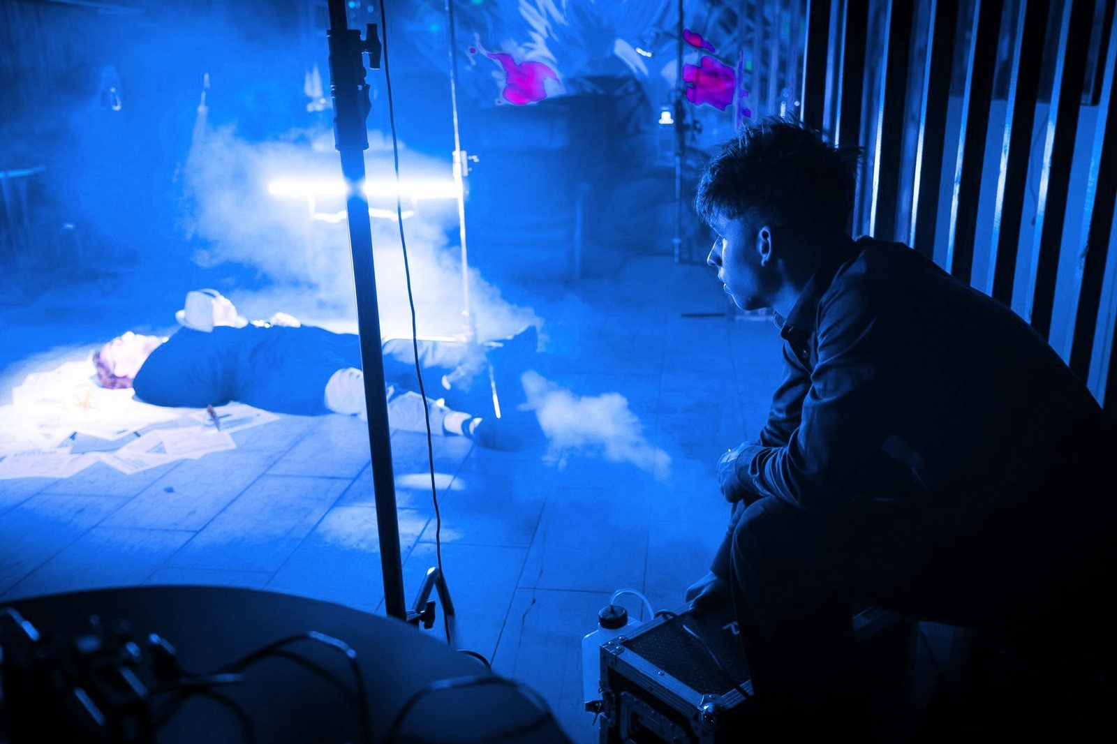 A crew member crouches by a smoke machine in blue light while an actor lies on the set behind a light stand.