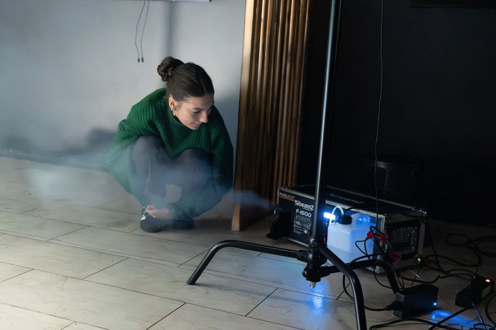 A crew member in a green sweater crouches by a smoke machine, spreading haze across the floor.