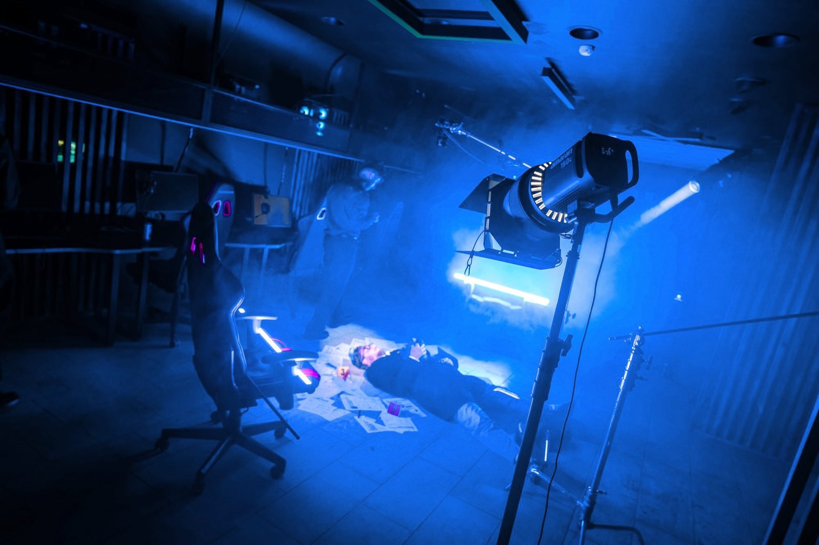 A spotlight on a stand lights a scene in blue haze, with an actor lying on the floor among scattered papers.